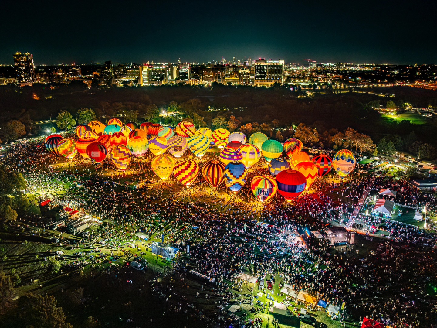 Aerial view of illuminated hot-air balloons, production areas, and a large crowd in Forest Park