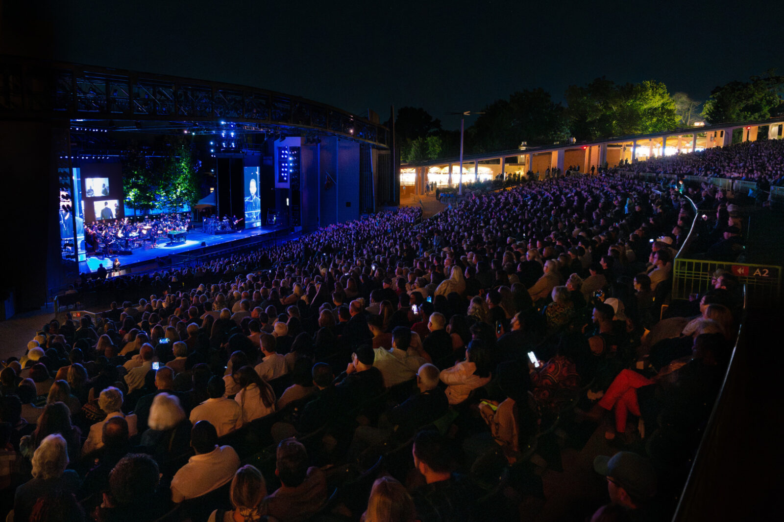 Wide nighttime view of the John Legend concert stage and full audience at The Muny