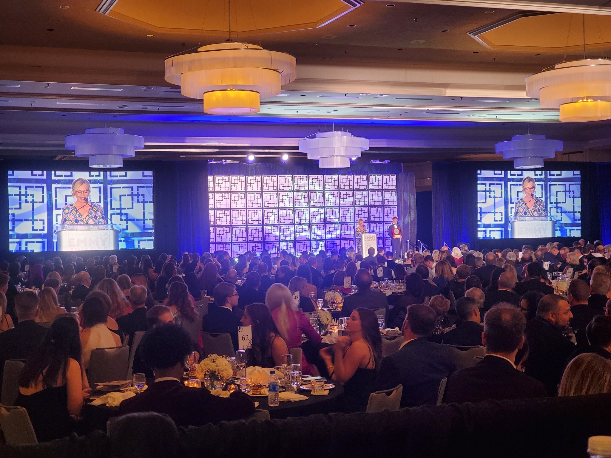 Wide banquet-room view of the Mid-America Emmy Awards stage, geometric scenic backdrop, video screens, and audience