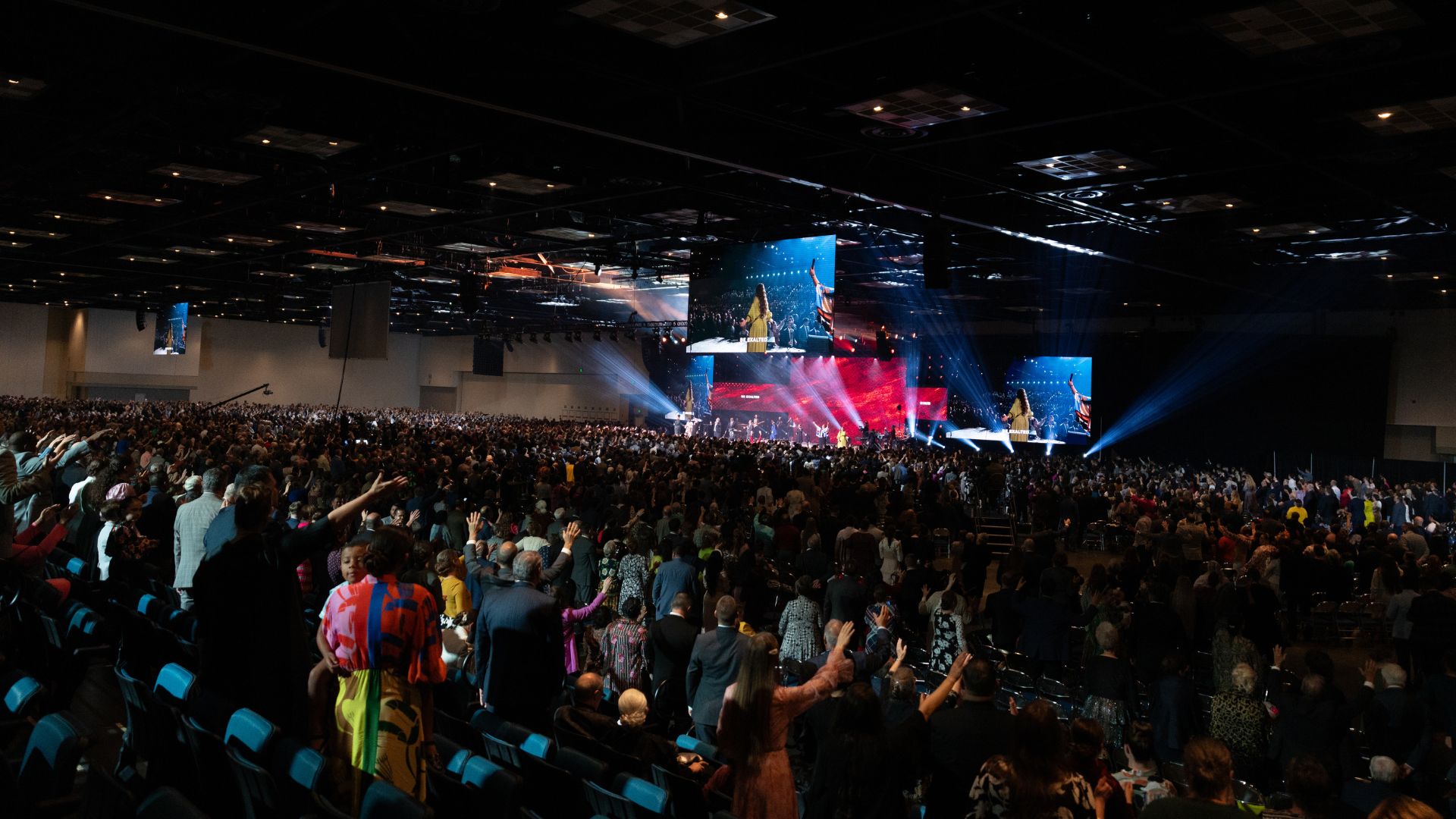 Arena-wide view of the United Pentecostal Church Annual Conference showing the audience, stage, video screens, and concert lighting